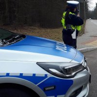 Policjant Wydziału Ruchu Drogowego, który mierzy prędkość  nadjeżdżających pojazdów, na zdjęciu widoczny jest także przód radiowozu