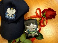 Na zdjęciu znajduje się czapka policyjna, etui na legitymacje służbową z odznaką policyjną i czerwona róża