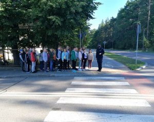 Uczniowie Szkoły Podstawowej nr 3 w Hajnówce wraz z funkcjonariuszką stoją przed przejściem dla pieszych. Policjanta tłumaczy dzieciom jak prawidłowo należy korzystać z przejścia dla pieszych.