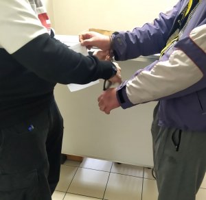 Funkcjonariusz z Wydziału Kryminalnego zakłada zatrzymanemu kajdanki na ręce trzymane z przodu