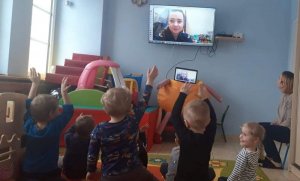 Zdjęcie przedstawia małe dzieci, które uczestniczą w spotkaniu online z policjantką. Na ekranie telewizora widać policjantkę.