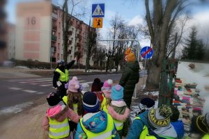 Zdjęcie przedstawia przedszkolaków w odblaskowych kamizelkach oraz policjantkę ruchu drogowego, która pokazuje dzieciom znak drogowy.