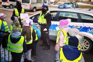 Zdjęcie przedstawia przedszkolaków w odblaskowych kamizelkach oraz policjantki, która pokazują dzieciom edukacyjne kolorowanki.