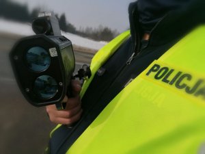 Zdjęcie przedstawia urządzenie do pomiarów prędkości, które w ręku trzyma policjant ruchu drogowego.