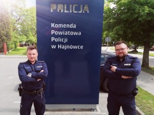 Zdjęcie przedstawia dzielnicowych. Policjanci stoją przy niebieskiej tablicy w napisem Policja, Komenda Powiatowa Policji w Hajnówce.