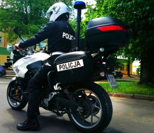 Zdjęcie przedstawia policjanta na motocyklu służbowym. Policjant ubrany jest w czarny kombinezon z białym napisem Policja. Motocykl jest w kolorze białym.