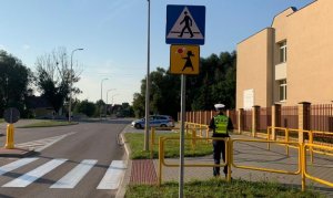 Zdjęcie przedstawia policjanta ruchu drogowego, stojącego przy barierkach ochronnych, w rejonie przejścia dla pieszych.