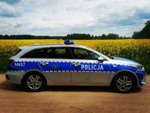Zdjęcie przedstawia oznakowany radiowóz, który jest ustawiony przy polu rzepaku.