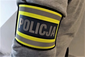 Ramię osoby ubranej w szarą bluzę, z założoną opaską z napisem &quot;Policja&quot;