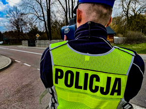 Na zdjęciu widoczny jest policjant drogówki, stojący tyłem. Dokonuje pomiaru prędkości przeznaczonym do tego urządzeniem.