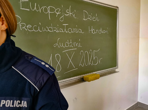 Tablica szkolna, na której widnieje napisany temat zajęć: Europejski Dzień Przeciwdziałania Handlowi Ludźmi. Na zdjęciu widać ramię policjanta.